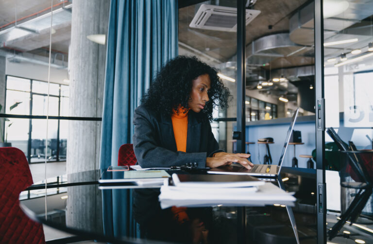Mulher sentada em uma mesa trabalha com ajuda de papeis e um notebook