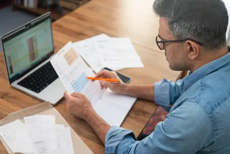 Homem analisando documentos financeiros e gráficos no notebook, avaliando dados de como abrir uma securitizadora