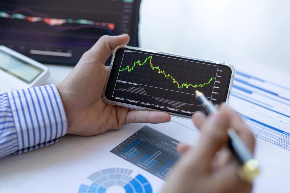 Mão segurando um smartphone com gráfico de mercado financeiro, representando a conversão de debêntures conversíveis em ações