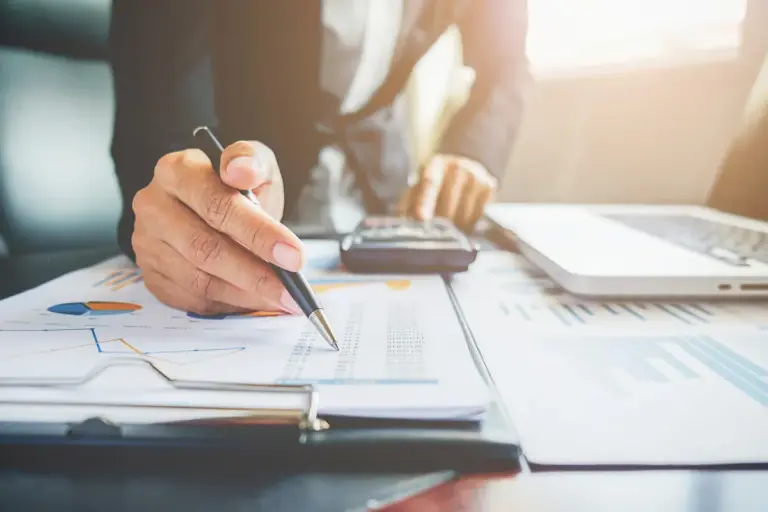 Executivo apontando caneta para planilha de dados financeiros com calculadora e laptop na mesa, analisando situação de cliente inadimplente