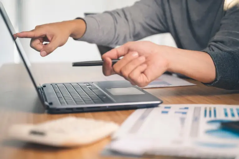 Mãos apontando para um notebook aberto ao lado de gráficos financeiros impressos, durante a análise de uma conta escrow