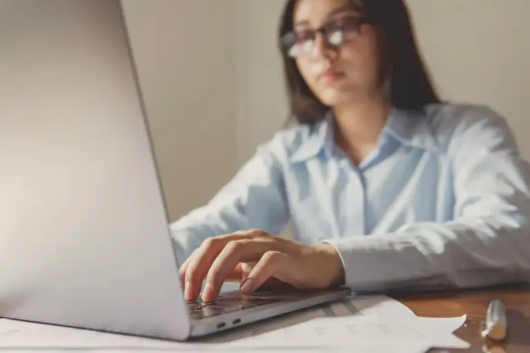 Mulher de óculos e camisa azul digita no notebook com papéis na mesa, emitindo uma duplicata escritural