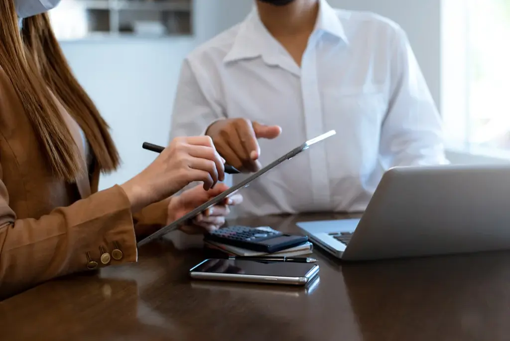 Duas pessoas analisando informações em um tablet, com calculadora, celular e notebook na mesa, discutindo o caso de um cliente inadimplente